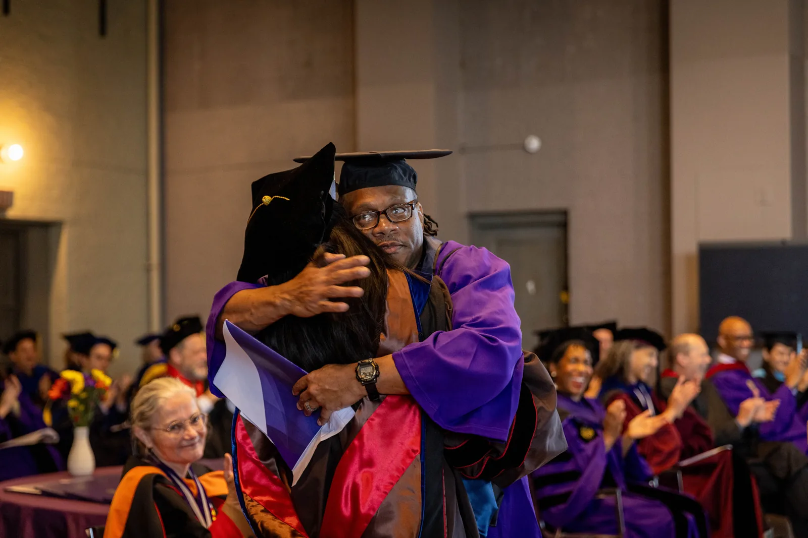 A man in a purple graduation gown and black mortarboard hugs a woman in academic regalia, with seated graduates and guests in the background.