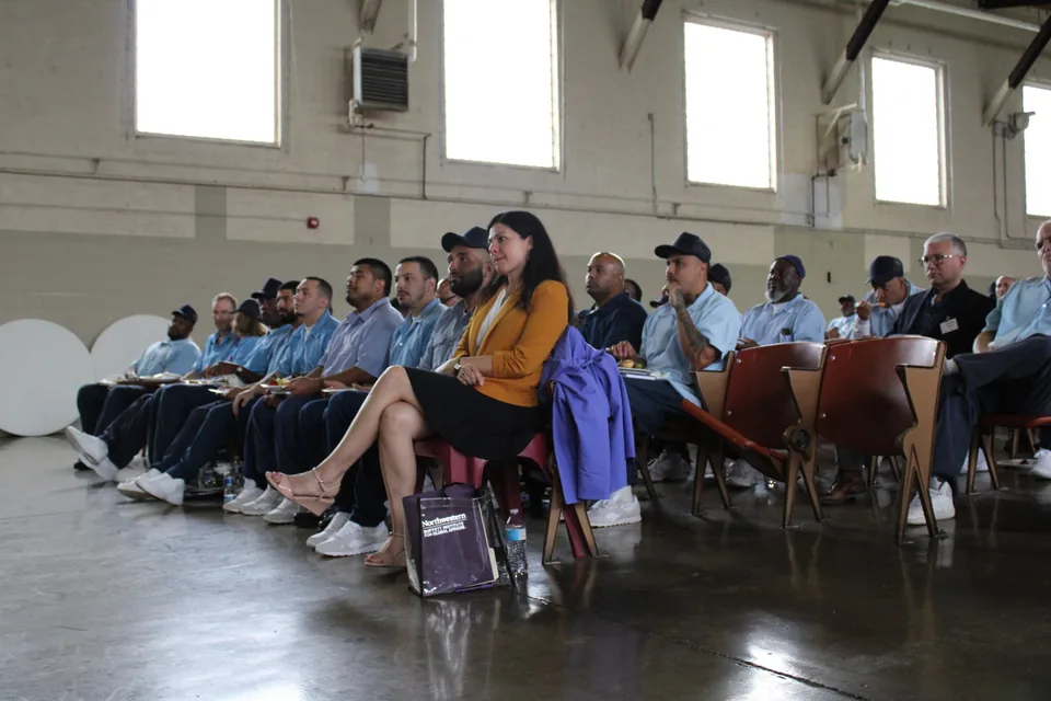 A woman in an orange top sits in the front row of a large audience of people in light blue prison uniforms seated in rows in a high-ceilinged hall.
