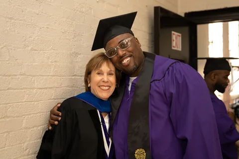 An older woman in academic regalia and a smiling man in a purple graduation gown and cap pose cheek to cheek.