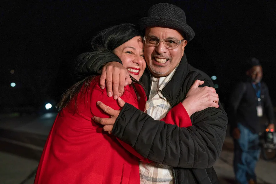 At night, a smiling man in glasses and a black hat embraces a smiling woman in a red coat, cheek to cheek.