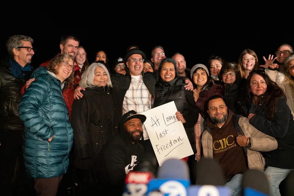 At night, a large group of smiling people crowds together around a man in a hat and plaid shirt; one woman holds a handwritten sign reading “At long last... Freedom,” and news microphones are in the foreground.
