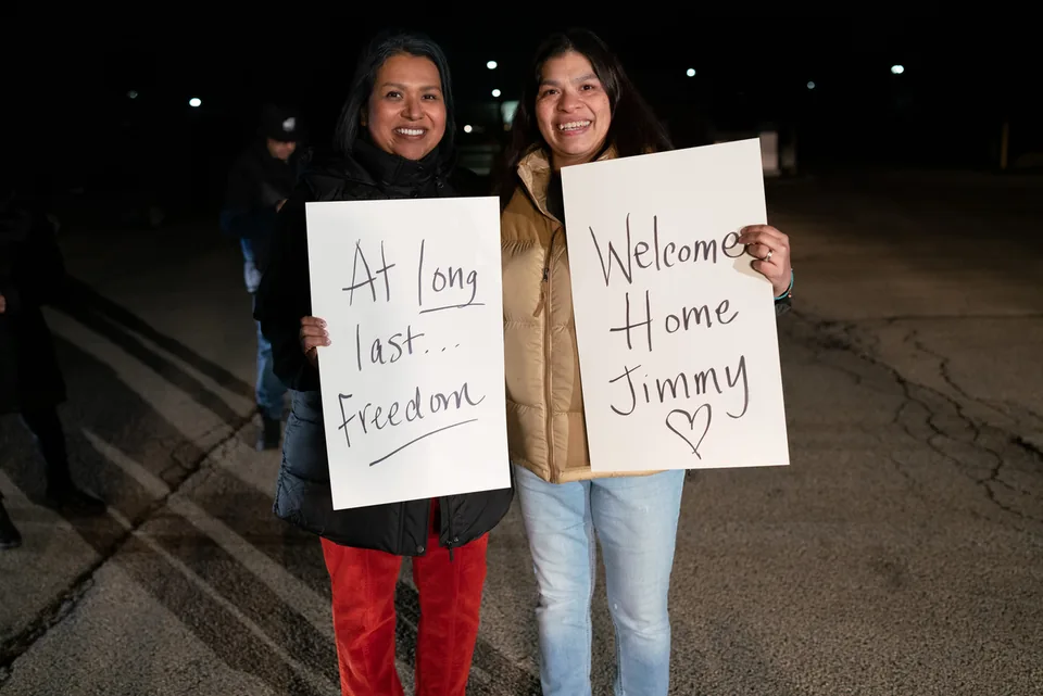 At night, two smiling women stand side by side holding handwritten signs reading “At long last... Freedom” and “Welcome Home Jimmy” with a heart.