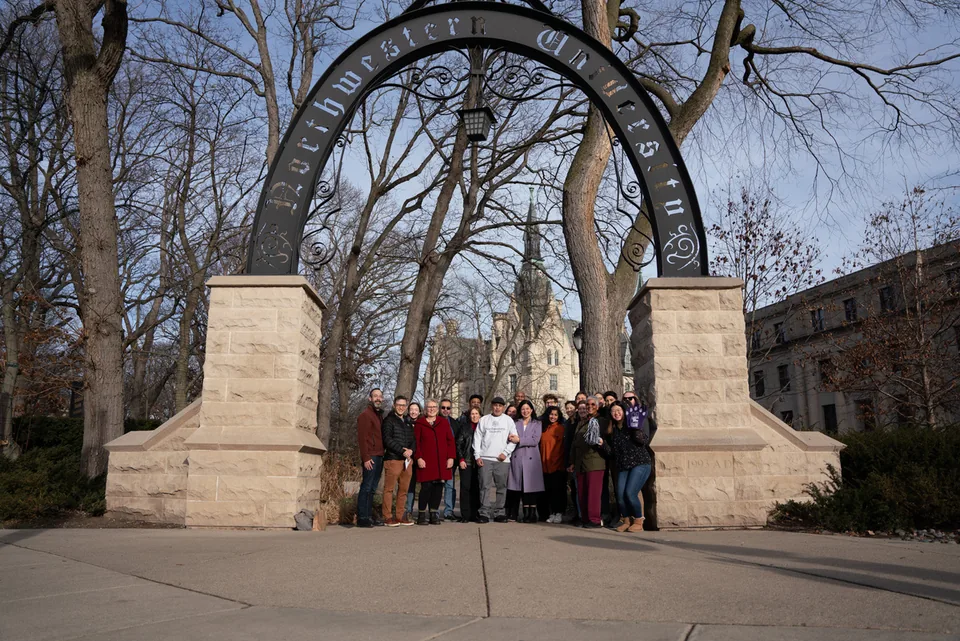 A large group of people poses together beneath a tall iron arch with “Northwestern University” lettering, flanked by stone pillars, with bare trees behind.