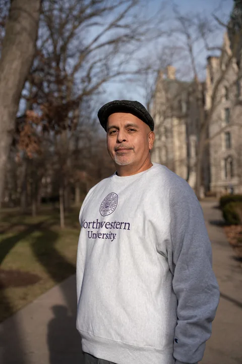 Outdoor portrait of a man in a dark flat cap and a gray Northwestern University sweatshirt standing on a tree-lined campus path.