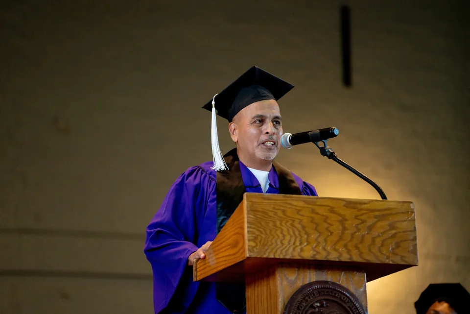A man in a purple graduation gown and black mortarboard with a white tassel speaks at a wooden podium microphone.