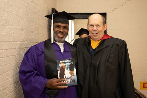 A smiling man in a purple graduation gown and mortarboard holds up a book while standing next to a smiling older man in black academic regalia.