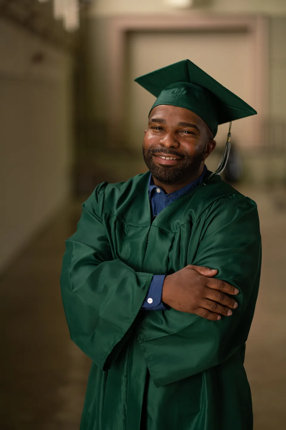 Portrait of a smiling man in a green graduation gown and mortarboard, arms crossed, against a dark background.