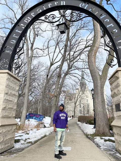A man in a purple Northwestern sweatshirt and light jeans stands on a snowy, tree-lined path between the stone pillars of an arched campus gate, with a large building in the background.