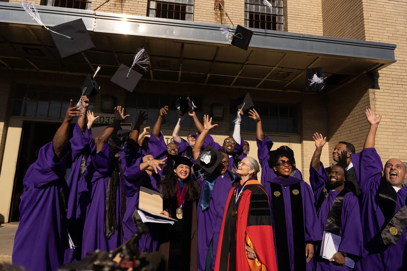 A group of graduates in purple gowns throw their mortarboards into the air and cheer, alongside smiling faculty members in academic regalia.