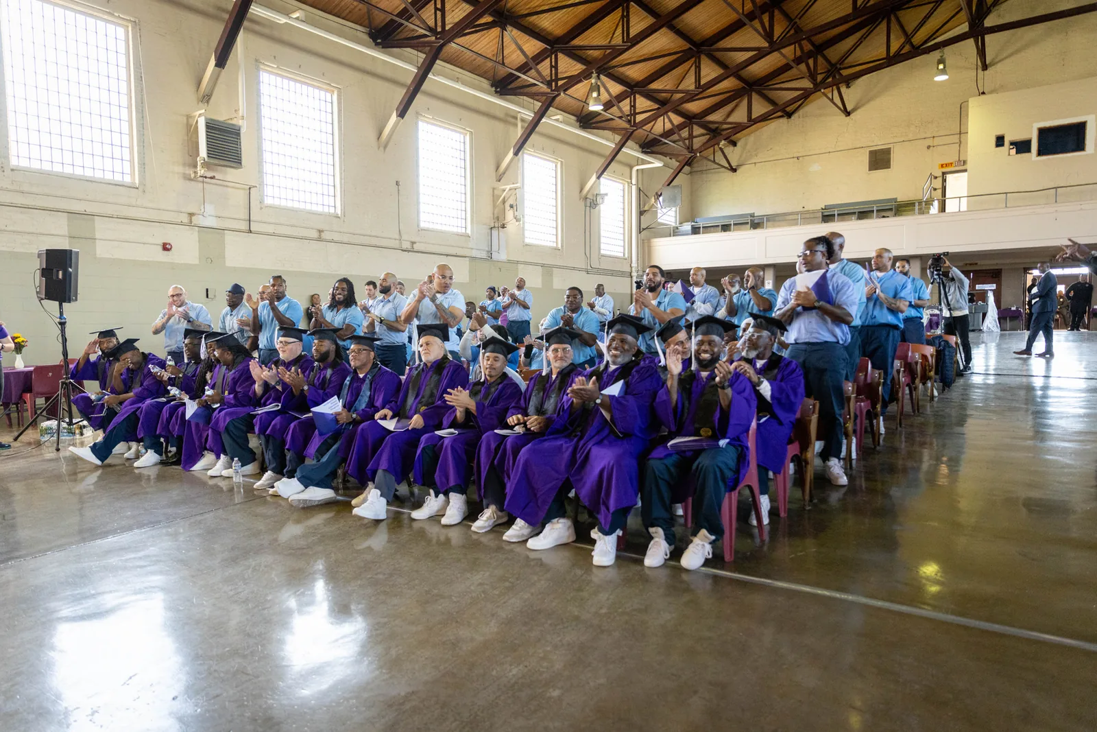 Wide view of a gymnasium-style hall where rows of graduates in purple gowns and mortarboards sit and applaud, with guests in light blue shirts seated behind them.