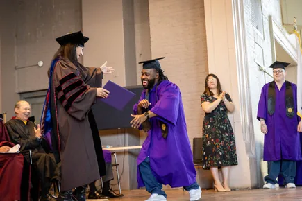 On a stage, a graduate in a purple gown and mortarboard shakes hands with a person in academic regalia, with others seated nearby.