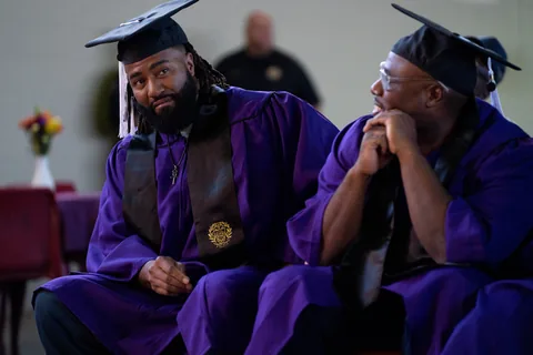 Two graduates in purple gowns and mortarboards sit side by side, one looking toward the other.