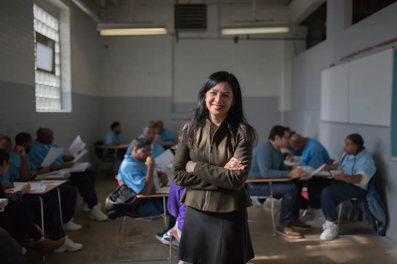 A smiling woman with long dark hair stands with arms crossed in a classroom where students in light blue uniforms sit at desks behind her.