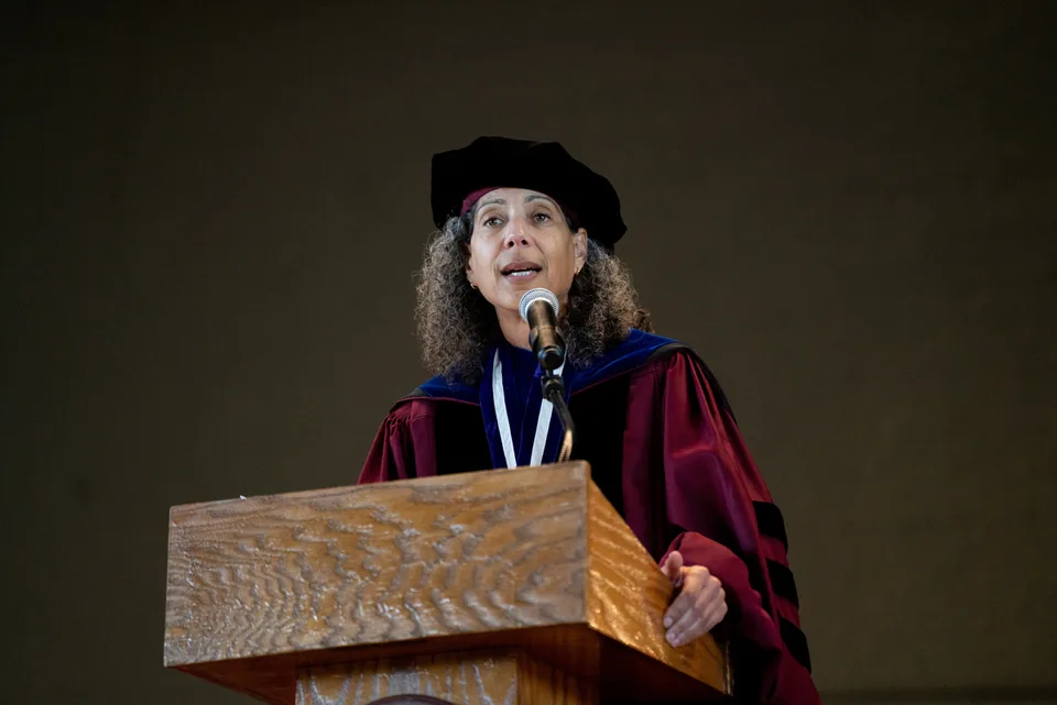A woman with curly hair wearing a black doctoral cap and red-and-blue academic robe speaks at a wooden podium microphone.
