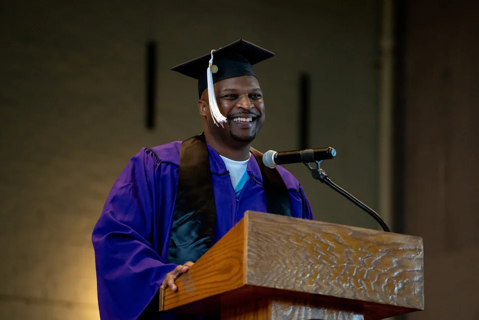 A smiling man in a purple graduation gown and black mortarboard with a white tassel stands at a wooden podium with a microphone.