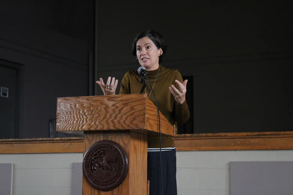 A woman in a brown sweater gestures while speaking at a wooden podium bearing a seal.