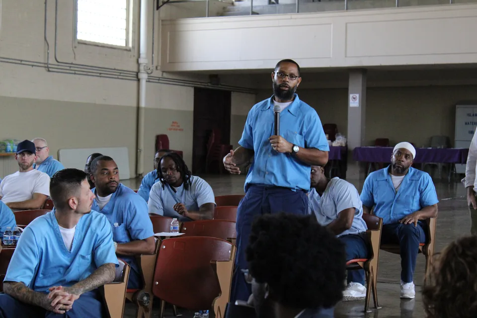 A man in a light blue uniform stands speaking among seated people, most in light blue uniforms, in a large hall.