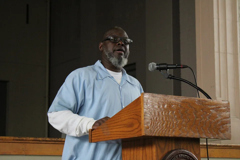 A man in a light blue uniform with glasses speaks at a wooden podium with a microphone; the accompanying quote identifies him as William Peeples.