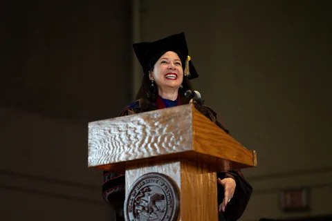 A smiling woman in academic regalia and cap speaks at a wooden podium bearing a state seal.