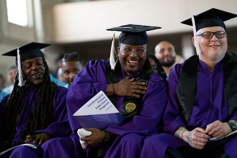 Graduates in purple gowns and mortarboards sit in a row smiling; the man in the center holds a program to his chest.