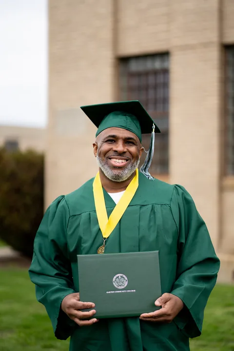 A smiling graduate in a green cap and gown with a yellow stole holds a diploma folder in front of a brick building.