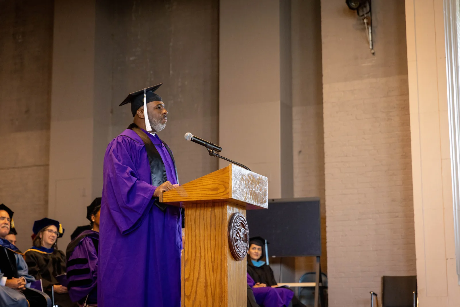 A graduate in a purple gown and mortarboard speaks into a microphone at a wooden podium.