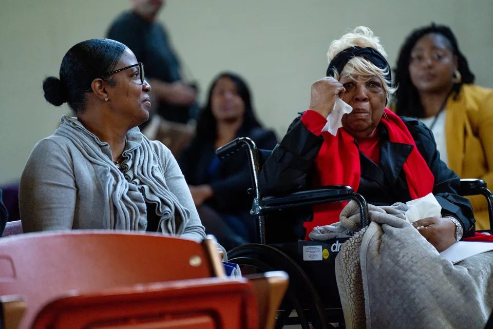 Women seated in the audience at a ceremony; an older woman in a red scarf wipes her eyes.