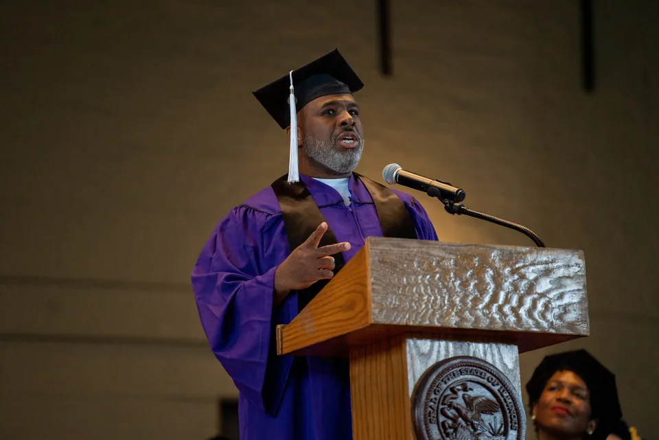 A graduate in a purple gown and mortarboard speaks and gestures at a podium with a microphone.