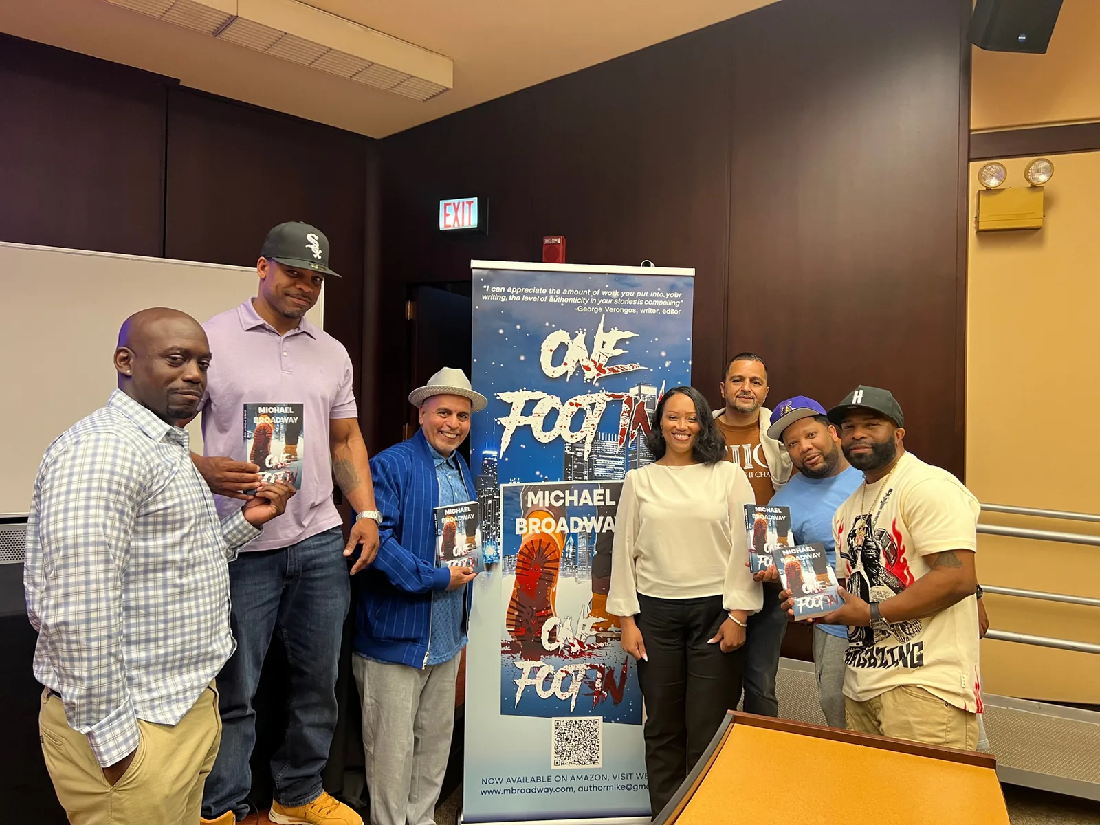 A group of people pose around a banner for the book “One Foot In” by Michael Broadway, several holding copies of the book.