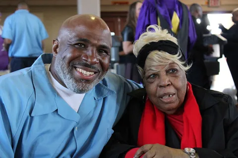 A smiling man in a light blue shirt poses cheek to cheek with an older woman wearing a black hat and a red scarf.