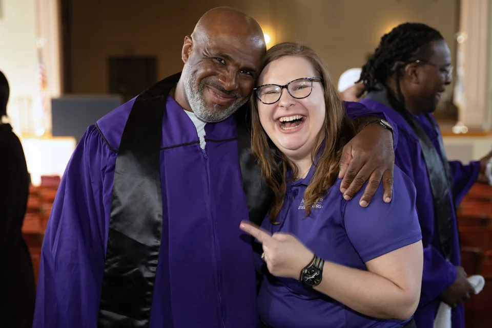 A smiling man in a purple gown puts his arm around a laughing woman in glasses and a purple shirt.