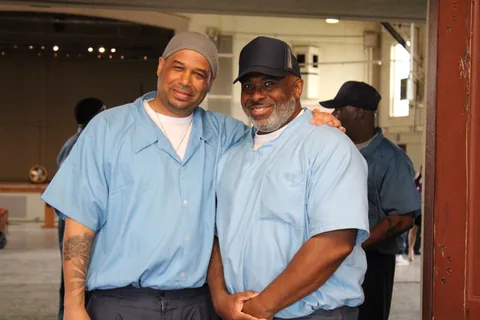 Two men in light blue uniforms stand with their arms around each other in a doorway.