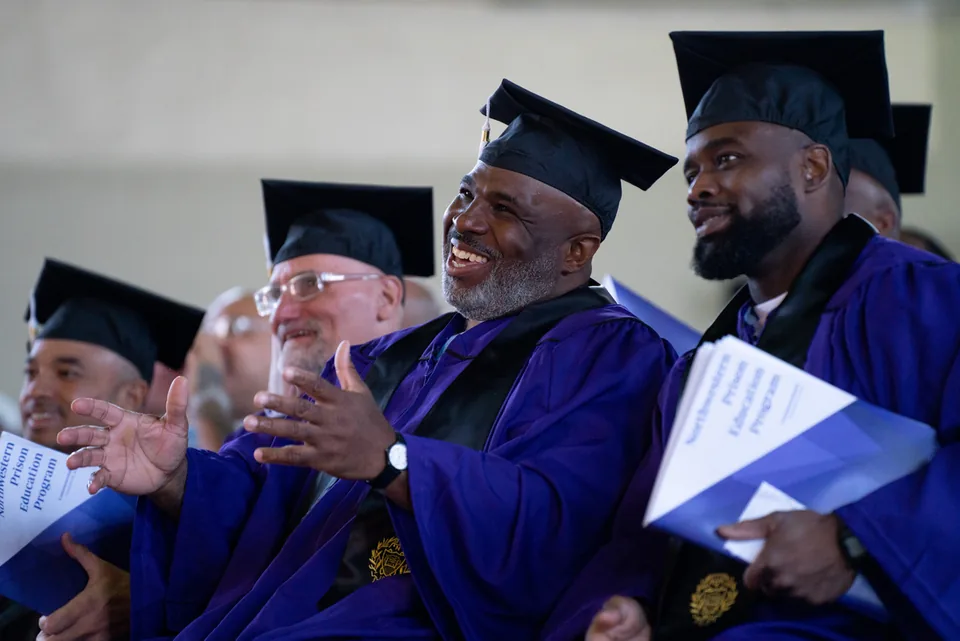A smiling graduate in a purple gown and mortarboard claps while seated among other graduates.