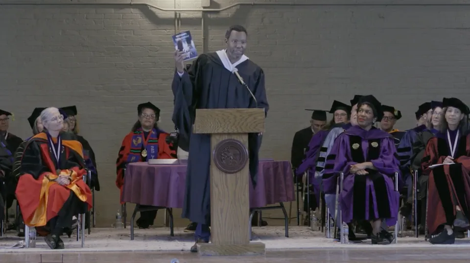 A graduate in a dark gown stands at a podium holding up a book.