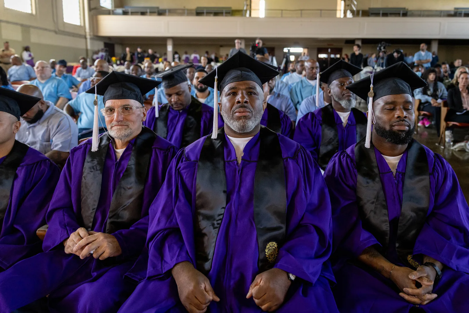 A graduate in a purple gown and mortarboard sits among other graduates, looking upward.
