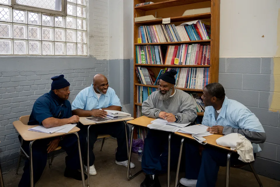 Four men sit at classroom desks talking, with a bookshelf full of binders behind them.