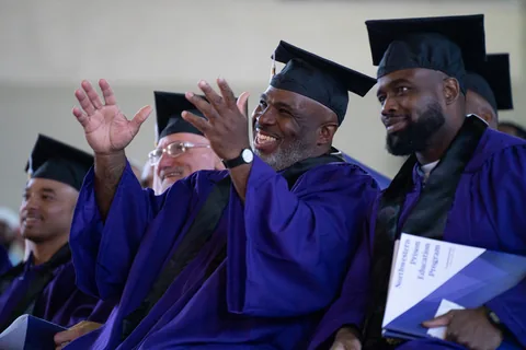 Graduates in purple gowns and mortarboards sit and clap during a ceremony.