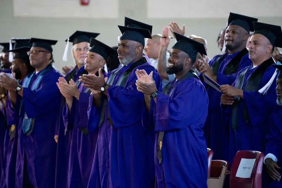 Graduates in purple gowns and mortarboards stand and clap.