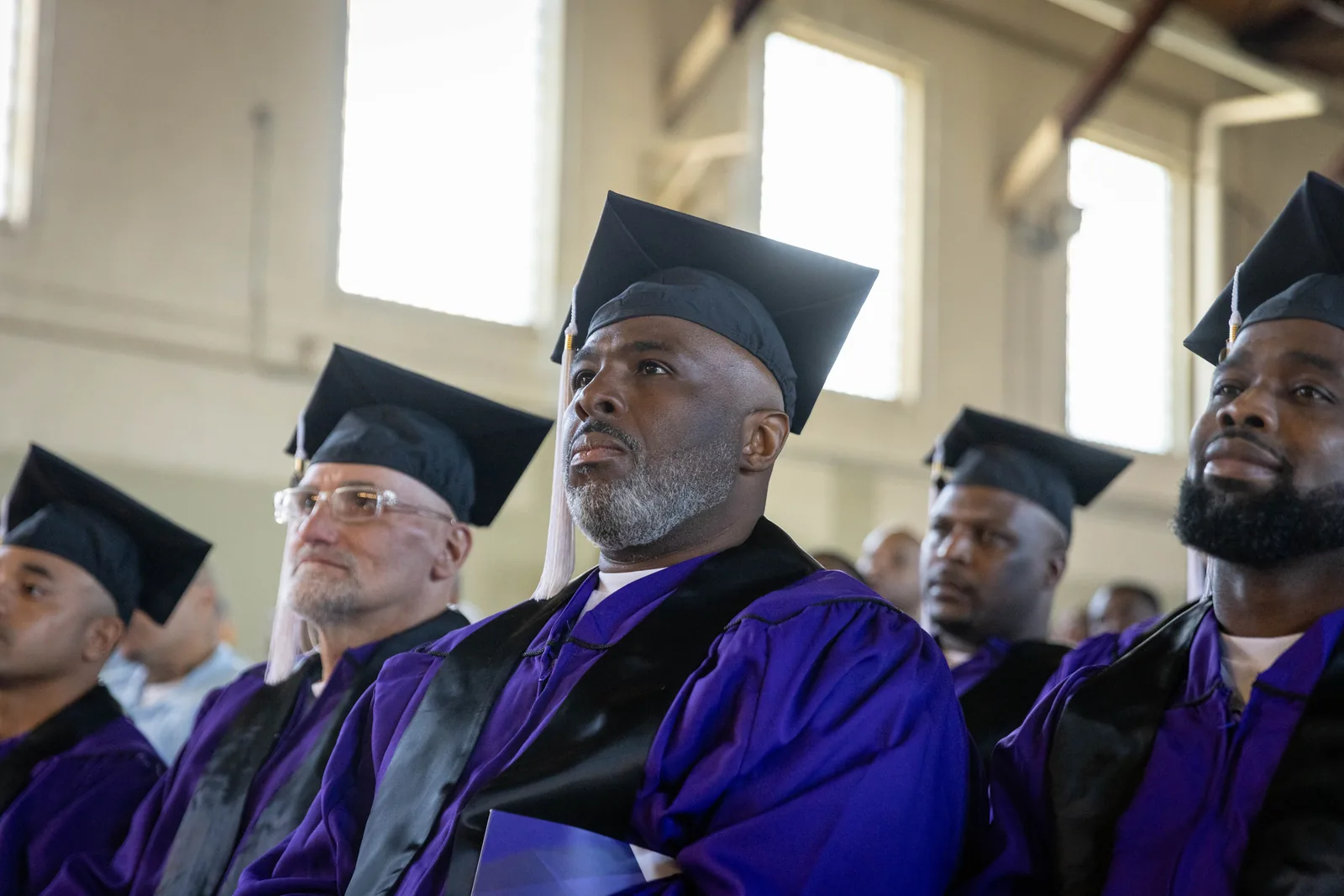 A graduate in a purple gown and mortarboard sits looking upward.