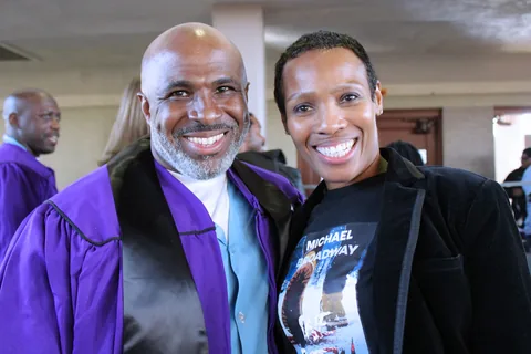 A smiling man in a purple graduation gown poses with a smiling woman whose T-shirt is printed with “Michael Broadway” and a book cover.