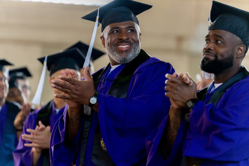 A smiling graduate in a purple gown and mortarboard claps his hands.