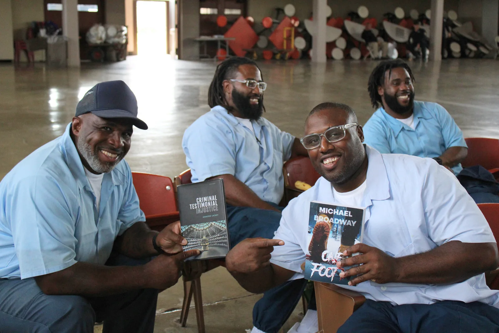 Three smiling men sit together; one holds up a copy of the book “One Foot In” by Michael Broadway and another holds a different book.