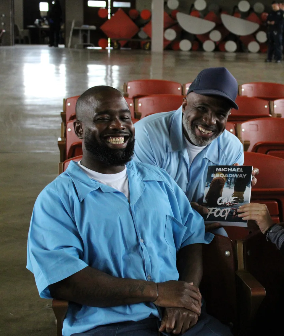 Two smiling men in light blue uniforms; one holds up a copy of the book “One Foot In” by Michael Broadway.