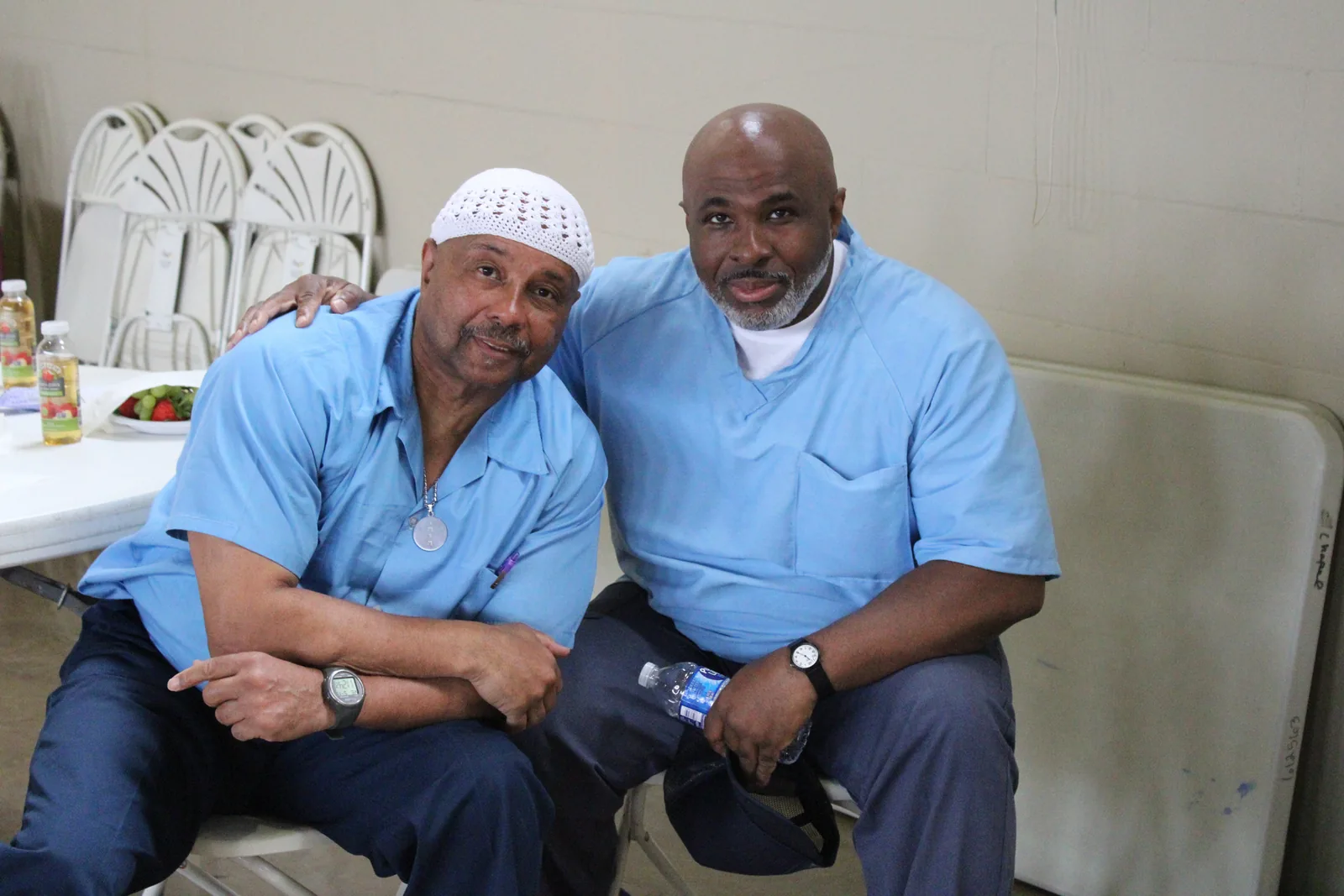 Two men in light blue shirts sit side by side, one with his arm around the other, both smiling.