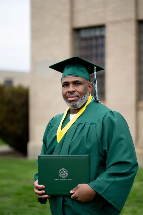 A graduate in a green cap and gown with a yellow stole holds a diploma folder in front of a brick building.