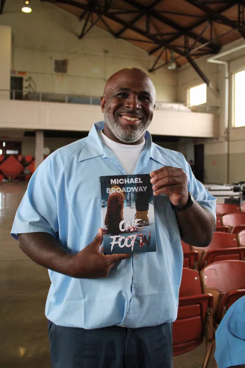 A smiling man in a light blue button-up shirt stands in a large room with rows of red seats, holding up a copy of the book One Foot In.