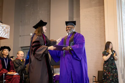 At a commencement ceremony, a smiling graduate in a purple gown and black cap shakes hands with a faculty member in academic regalia and a black cap while handing over a purple diploma folder; seated faculty and a woman clapping are in the background.
