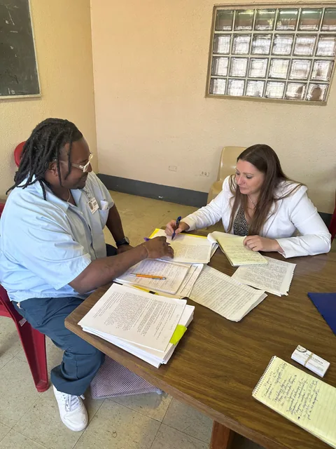 A man in a light blue shirt writes on papers at a table covered with documents while a woman in a white jacket sits across from him, leaning in.