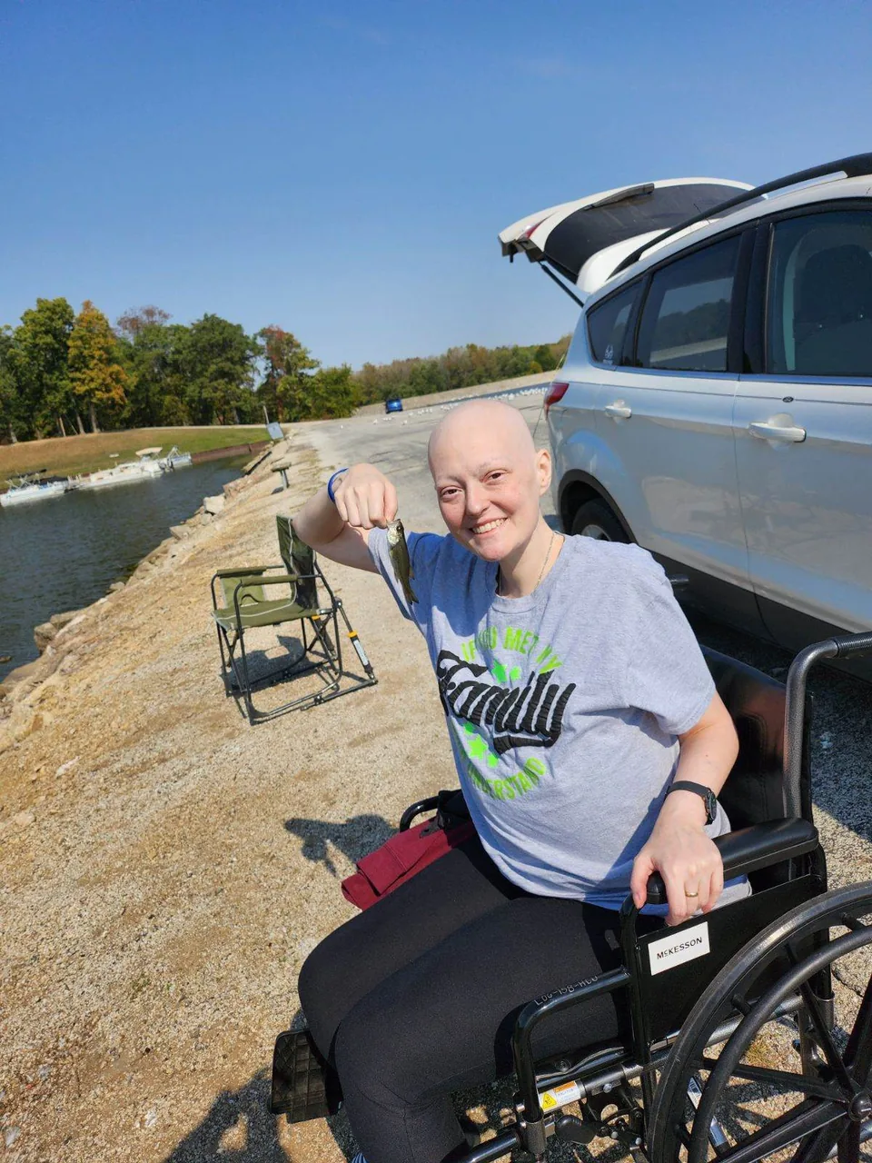 A smiling woman with a shaved head, wearing a gray T-shirt and seated in a wheelchair, holds up a small fish beside a lake, with a silver SUV and a folding chair behind her.