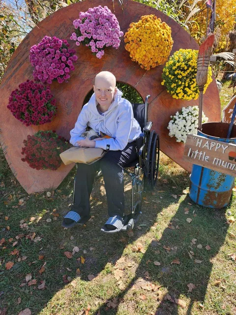 A smiling woman with a shaved head, in a light hooded sweatshirt and slides, sits in a wheelchair in front of a decorative display of potted mums on fallen leaves.